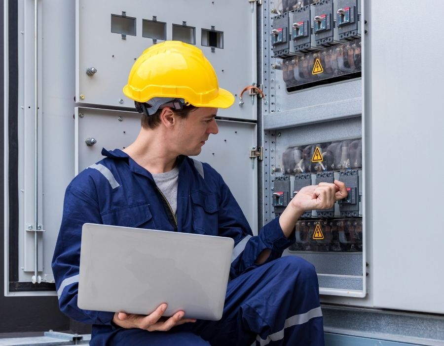 Technician wearing a hard hat and holding a laptop while inspecting industrial equipment, representing connected sales, service, and operational data.
